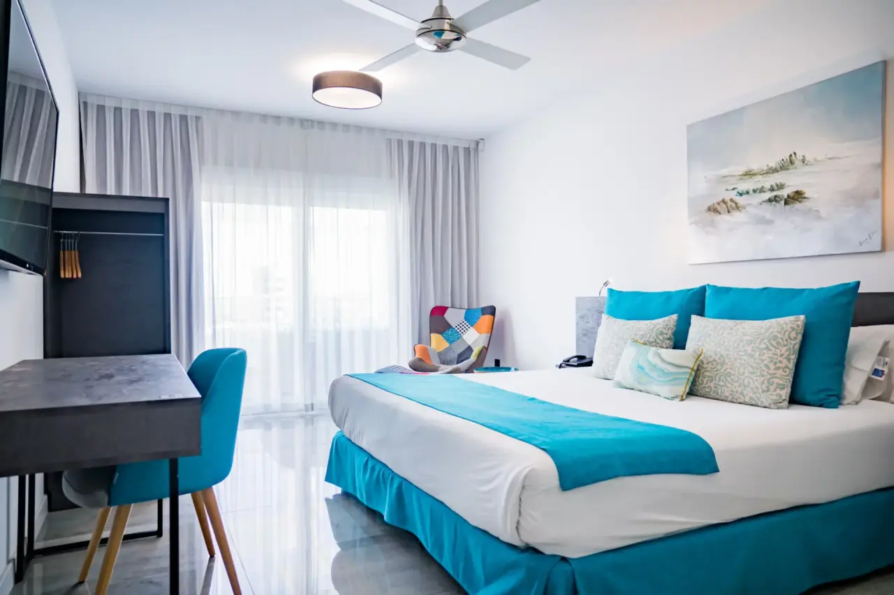 Habitación de hotel con cama grande vestida en blanco y turquesa, escritorio con silla azul, televisor en la pared