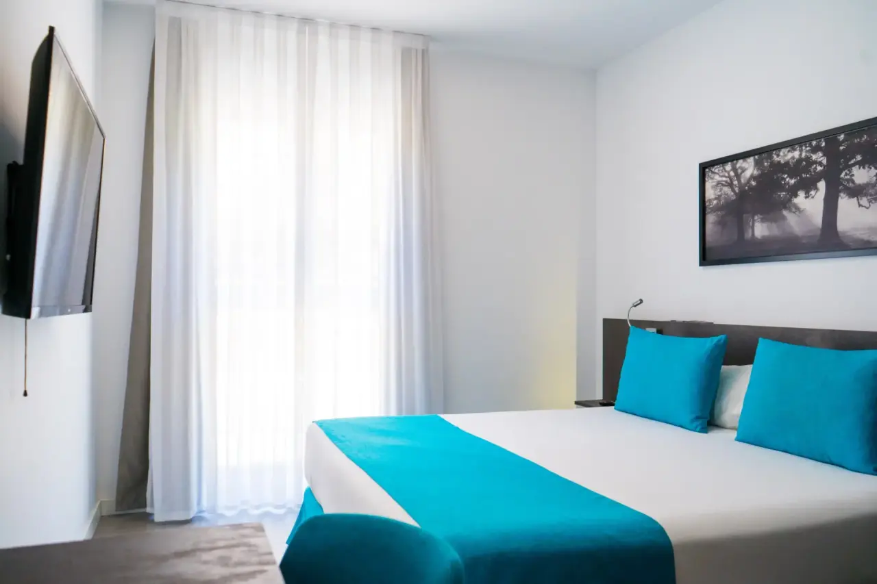 Habitación de hotel con cama doble vestida con sábanas blancas y una franja turquesa, dos almohadas turquesa, cabecero