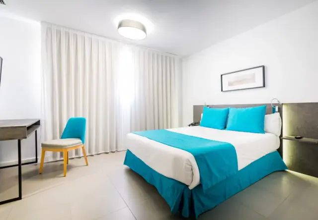 Habitación de hotel con cama doble vestida en blanco y turquesa, cabecero de madera, escritorio con silla azul