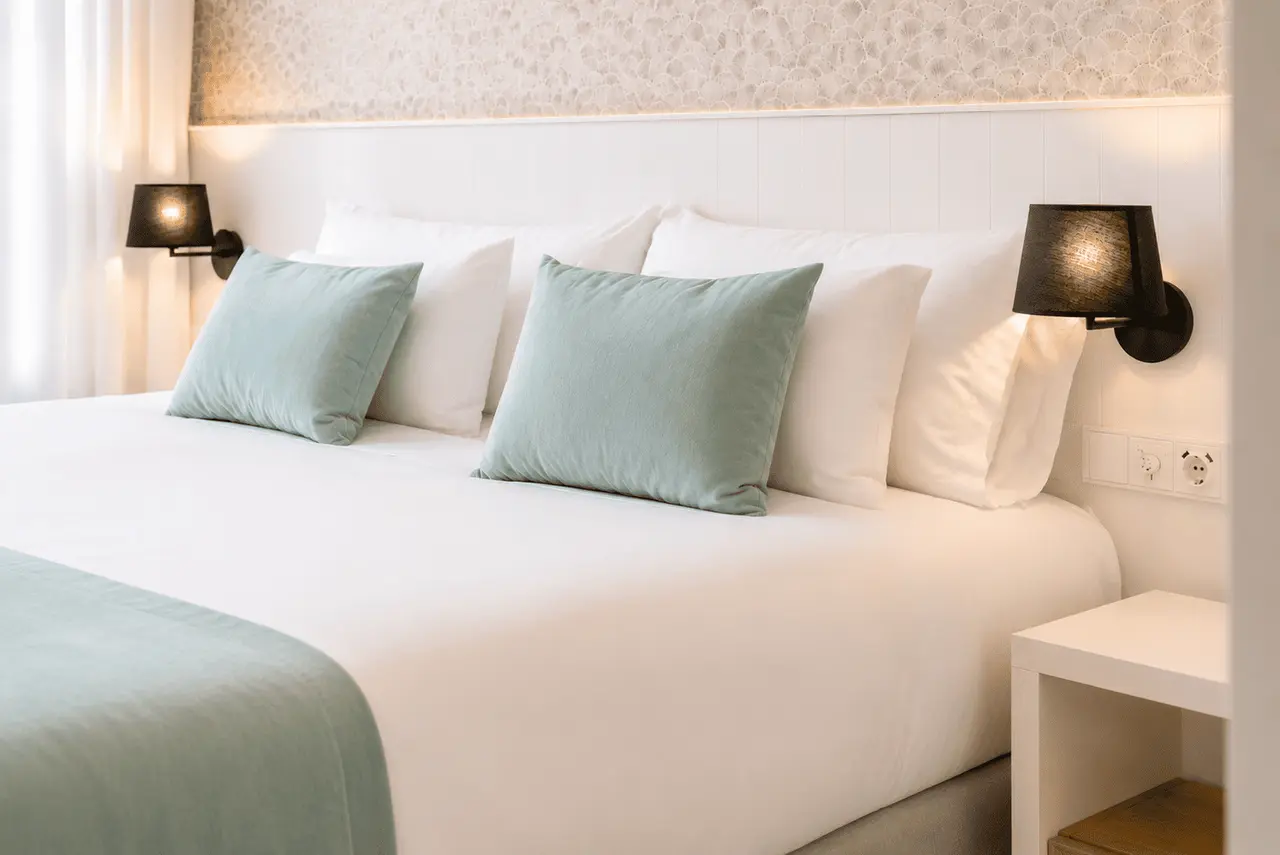 Cama doble en una habitación de hotel con sábanas blancas, cojines en tonos azul claro y blancos, cabecero blanco