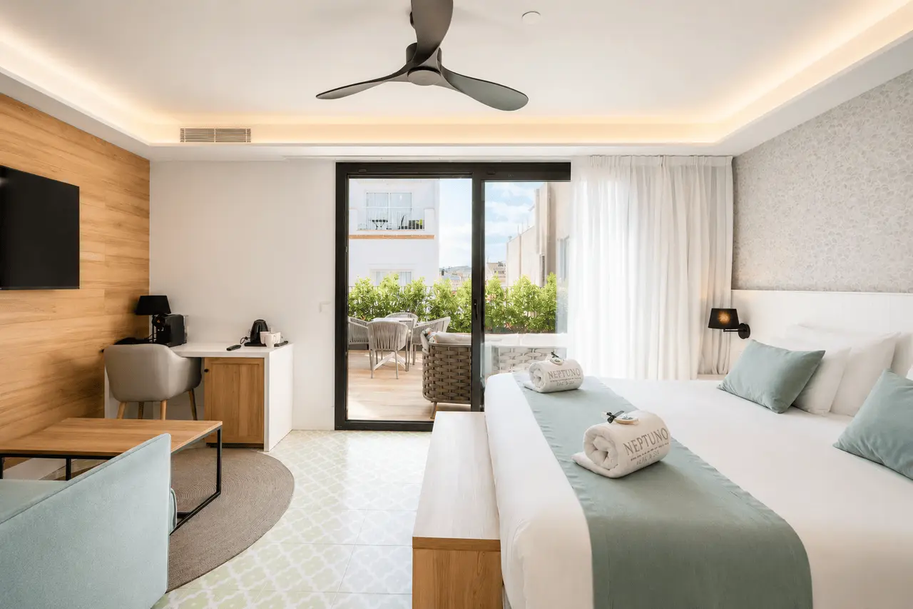 Vista de una suite de hotel con cama doble grande vestida de blanco y turquesa, toallas enrolladas sobre la colcha
