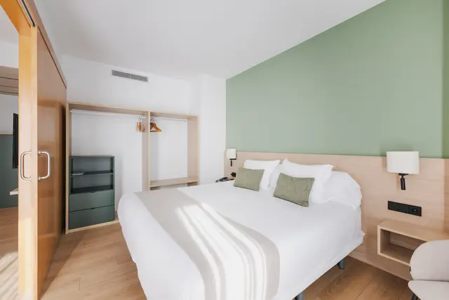 Habitación de hotel con cama doble grande vestida de blanco, cabecero de madera clara, dos almohadas verdes, mesitas