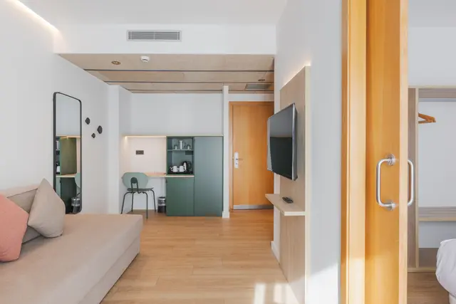 Vista de una suite moderna con sofá beige, espejo de cuerpo entero, zona de cocina con armarios verdes, televisor