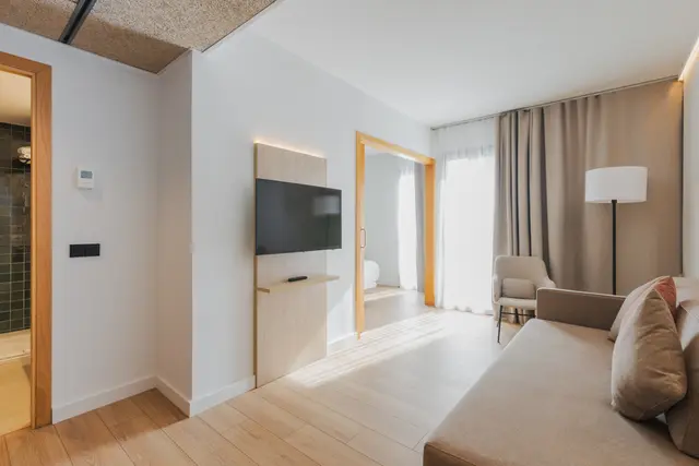 Interior de una suite con paredes blancas, suelo de madera clara, sofá beige en primer plano, televisor montado