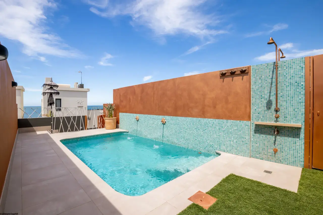 Piscina privada rectangular con agua azul en una terraza exterior rodeada de paredes con azulejos y césped artificial.