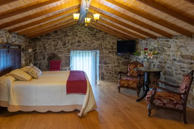 Interior de una suite con techo abuhardillado de madera, paredes de piedra, cama grande con colcha blanca y roja
