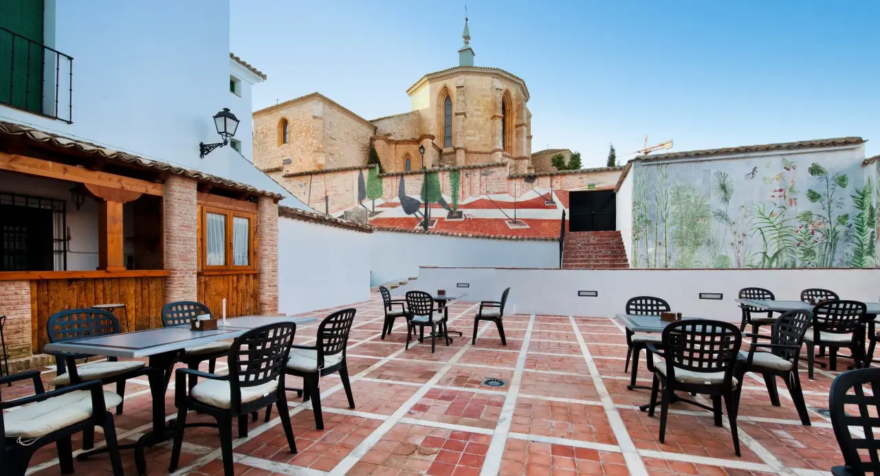 Terraza exterior con suelo de baldosas rojas, mesas y sillas de exterior dispuestas, rodeada de edificios blancos