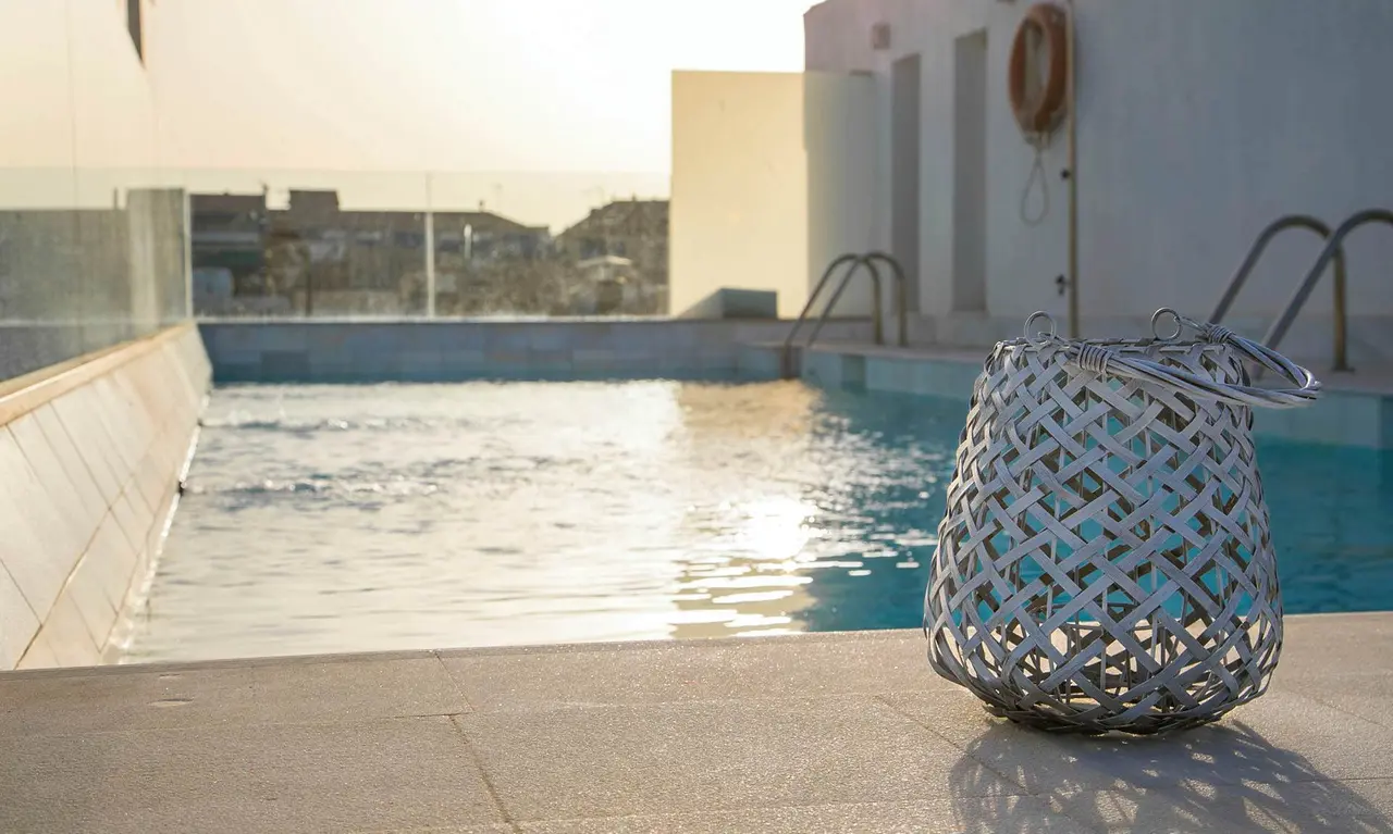 Vista de una piscina exterior con agua en movimiento y reflejos del sol, en primer plano un cesto de mimbre decorativo