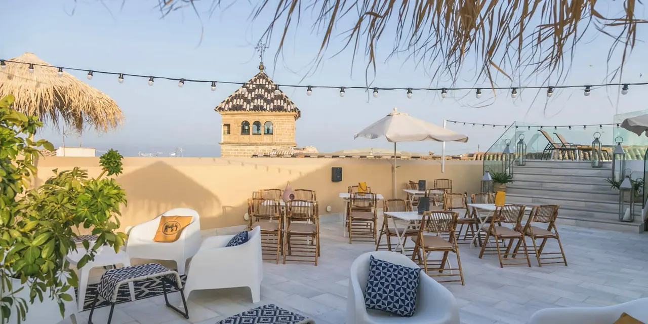 Terraza exterior de un hotel con suelo de piedra clara, muebles de exterior blancos y de madera, mesas, sillas