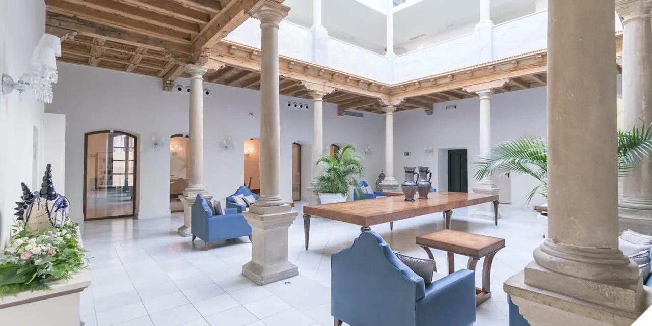 Interior de un amplio salón con suelo de baldosas blancas, columnas de piedra, techo de vigas de madera y claraboya.