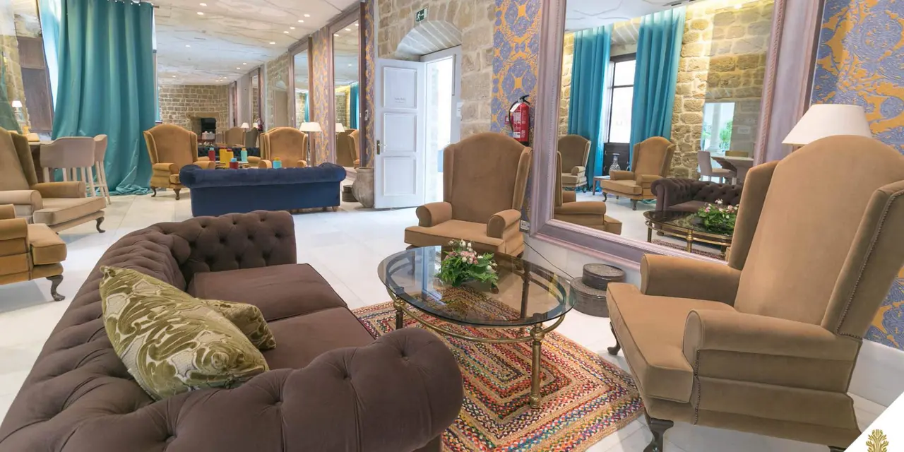 Vista de una sala de estar en una suite con sofá marrón capitoné en primer plano, sillones beige, mesa de centro