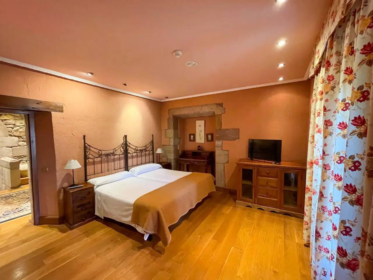 Vista de una suite de hotel con una cama de matrimonio con cabecero de forja, mesitas de noche, un mueble de madera