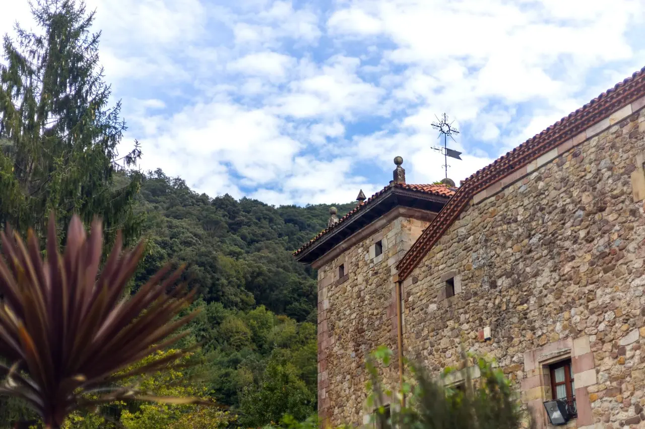 Vista exterior de un edificio de piedra con tejado de teja, rodeado de vegetación y un cielo nublado.