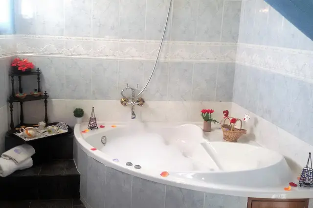 Baño con bañera de hidromasaje blanca de esquina, llena de agua y espuma, con pétalos de rosa flotando.