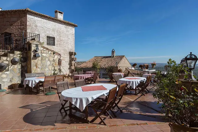 Terraza exterior de un edificio de piedra con mesas y sillas de madera dispuestas para comer, bajo un cielo azul