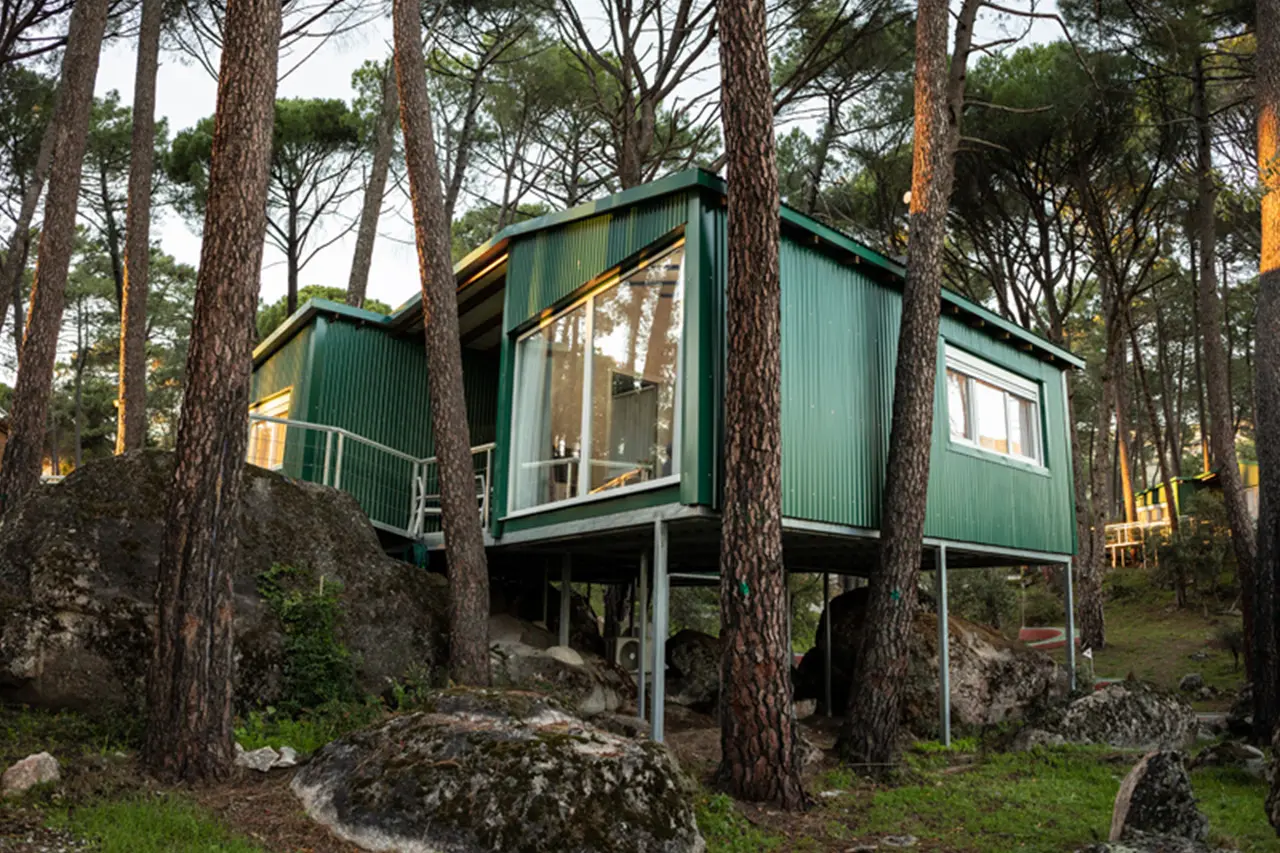 Vista exterior de una cabaña elevada de color verde con grandes ventanales, construida entre pinos altos y rocas