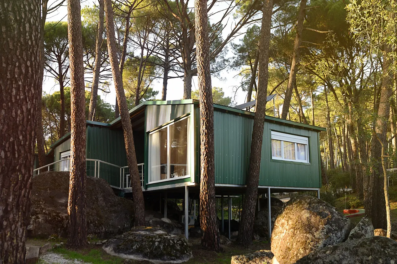 Vista exterior de una cabaña verde elevada sobre pilotes en un bosque de pinos, con grandes rocas en primer plano