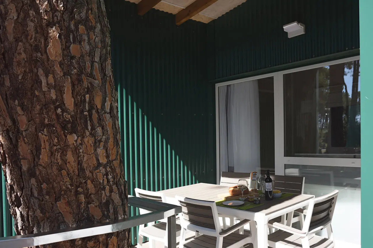 Terraza exterior con mesa y sillas blancas, junto a una pared verde y una gran ventana corredera.