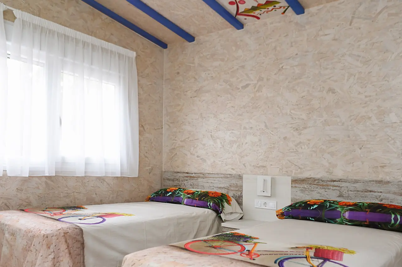 Habitación con dos camas individuales, paredes de paneles de madera clara, techo con vigas azules y decoración