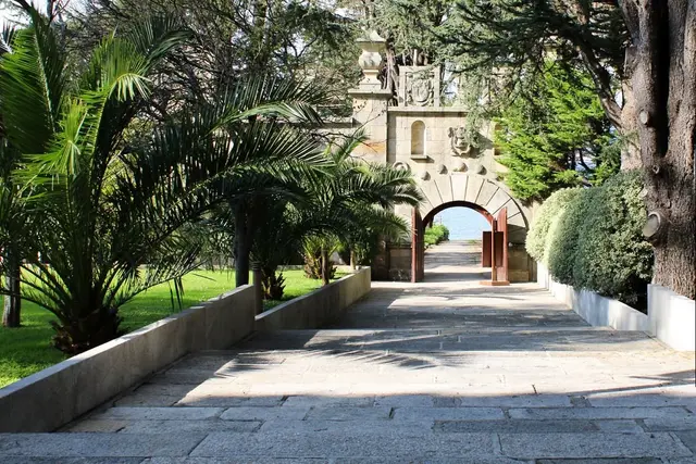 Camino empedrado rodeado de palmeras y vegetación que conduce a un arco de piedra con vistas al mar.