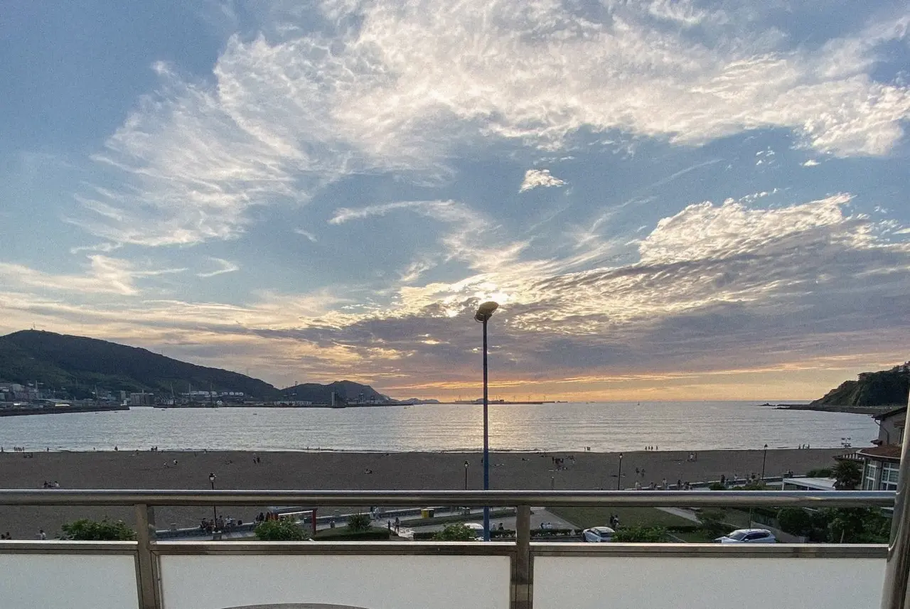 Vista desde un balcón hacia una playa extensa al atardecer, con el mar, nubes iluminadas y colinas al fondo.