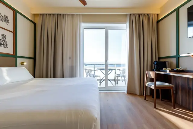 Vista de una junior suite con cama grande vestida de blanco, escritorio de madera con silla, puerta de cristal abierta a