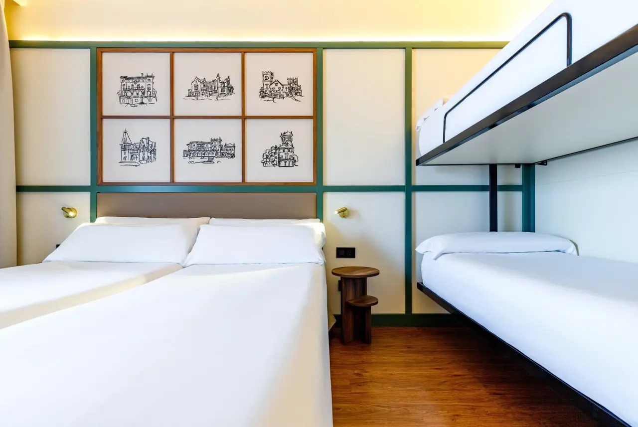 Habitación con dos camas individuales, una litera superior, cabecero con paneles verdes y seis cuadros de edificios