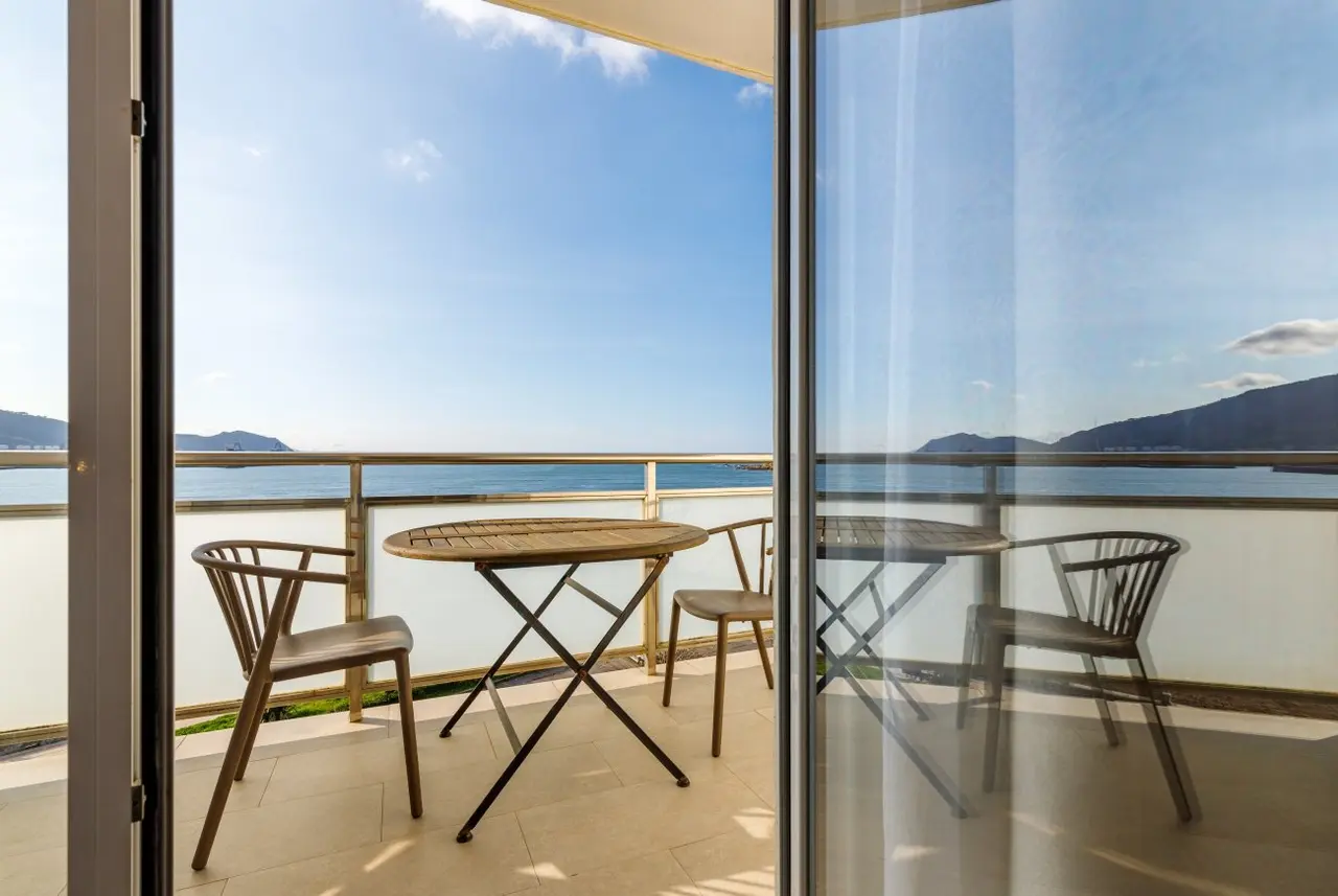 Vista desde el interior de una habitación hacia una terraza con mesa y sillas de madera, que da al mar y montañas