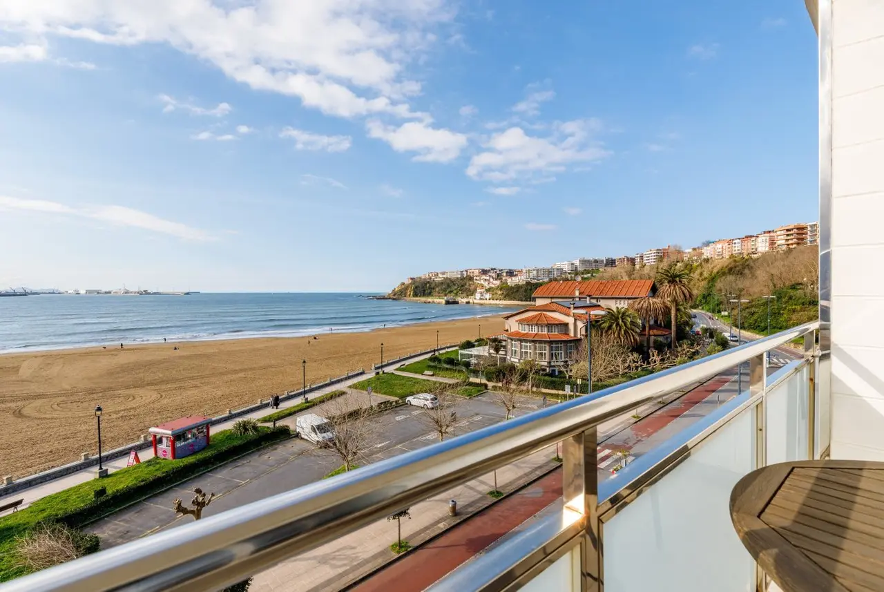Vista desde el balcón de la habitación hacia la playa, el mar y la ciudad costera bajo un cielo azul con nubes.