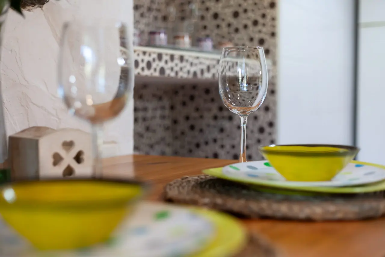 Mesa de madera con vajilla amarilla, cuencos, platos y copas de vino de cristal, sobre un salvamanteles de mimbre.