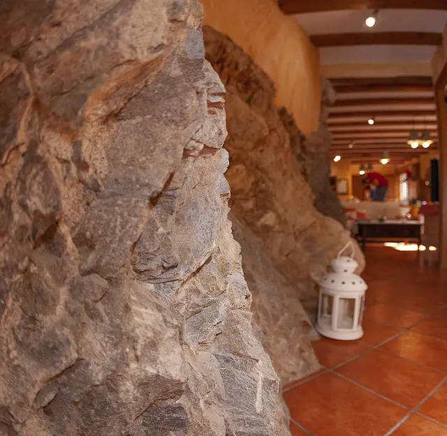 Interior de una casa rural con una pared de piedra irregular en primer plano, suelo de terracota y una linterna blanca