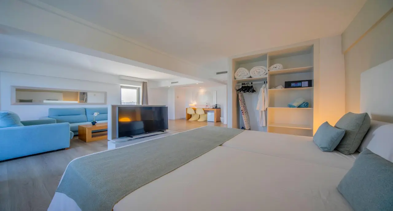 Vista de una junior suite superior con cama grande en primer plano, sofá azul, televisor, armario abierto con ropa