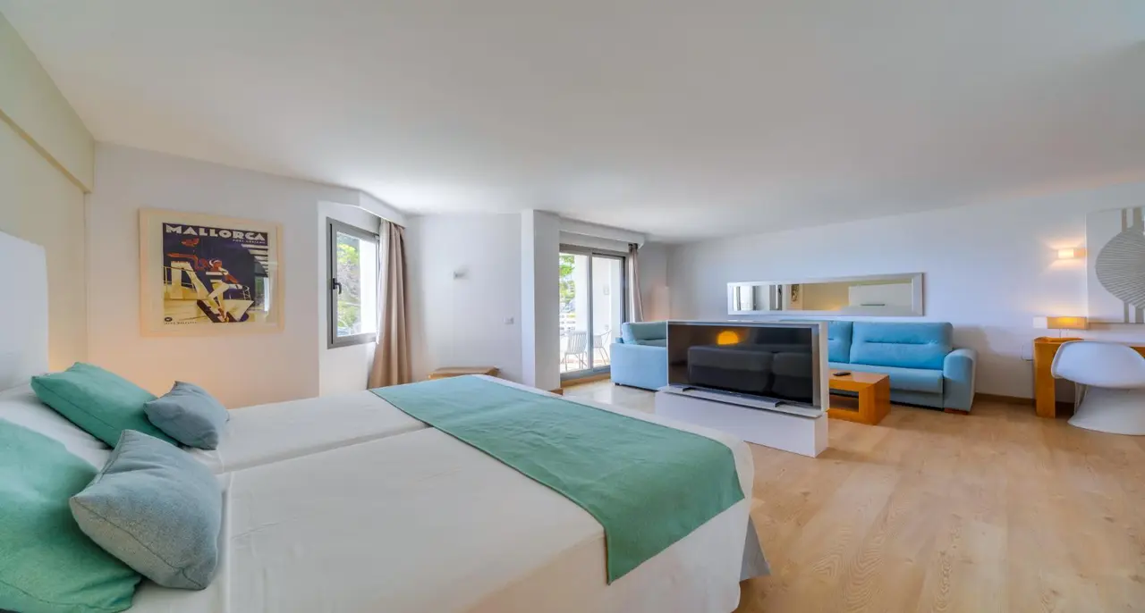 Vista de una junior suite con cama doble grande vestida de blanco y verde, sofá azul en la zona de estar, televisor