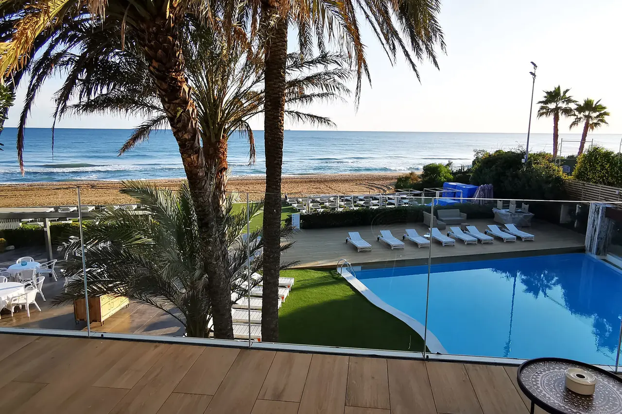 Vista desde una terraza con suelo de madera hacia una piscina rectangular de agua azul rodeada de césped y tumbonas