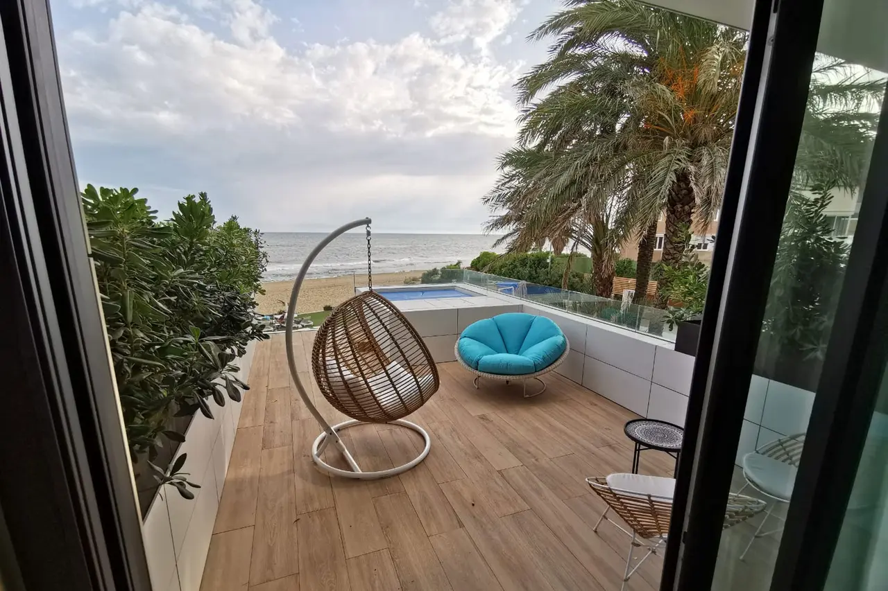 Vista desde el interior hacia una terraza con suelo de madera, una piscina rectangular privada, un sillón colgante