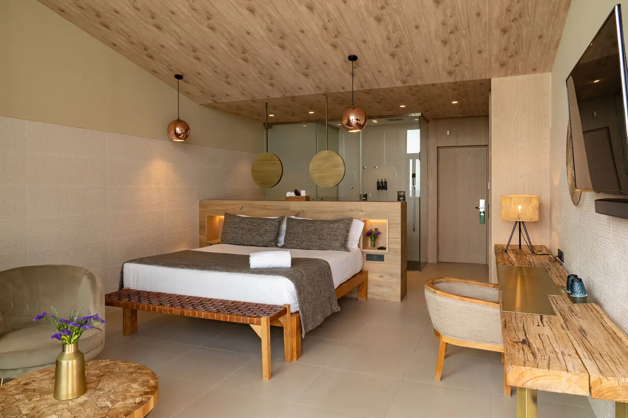 Habitación de suite con cama king size vestida con sábanas blancas y colcha gris, cabecero de madera iluminado