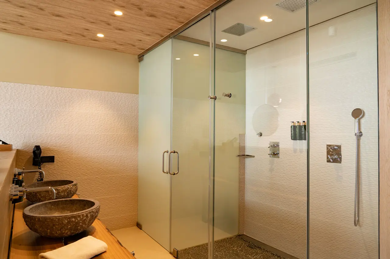 Baño interior con dos lavabos de piedra sobre encimera de madera, grifería negra, espejo y ducha cerrada con mampara