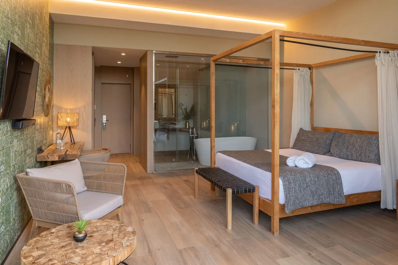 Interior de una suite con cama de dosel de madera, sillón de mimbre, mesa de centro redonda, televisor en la pared