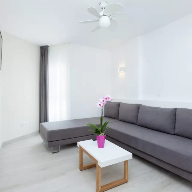 Interior de una suite con sofá gris en L, mesa de centro blanca con base de madera y una orquídea en maceta rosa