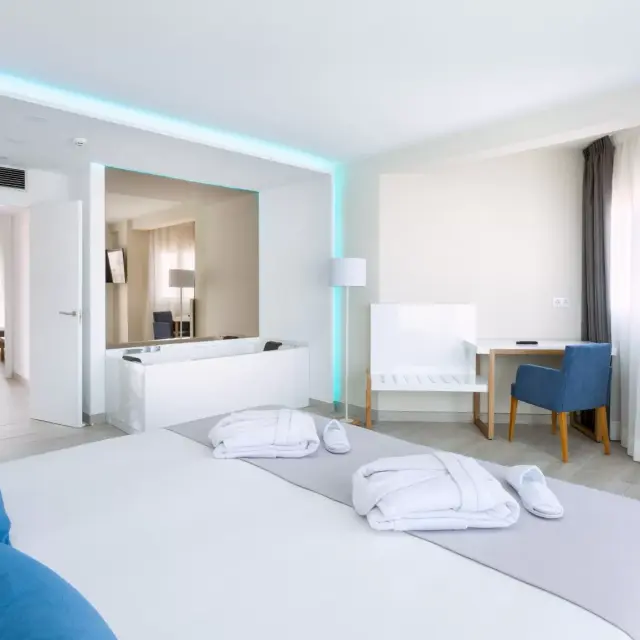 Interior de una suite con una bañera de hidromasaje blanca rectangular visible en un espacio abierto al fondo, cama