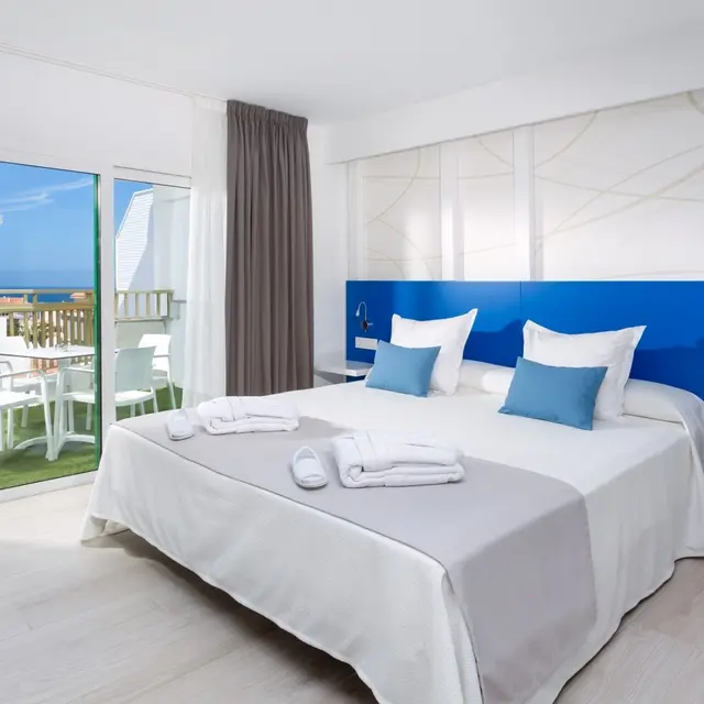 Habitación de hotel con cama doble grande vestida de blanco y azul, cabecero azul, toallas y zapatillas colocadas