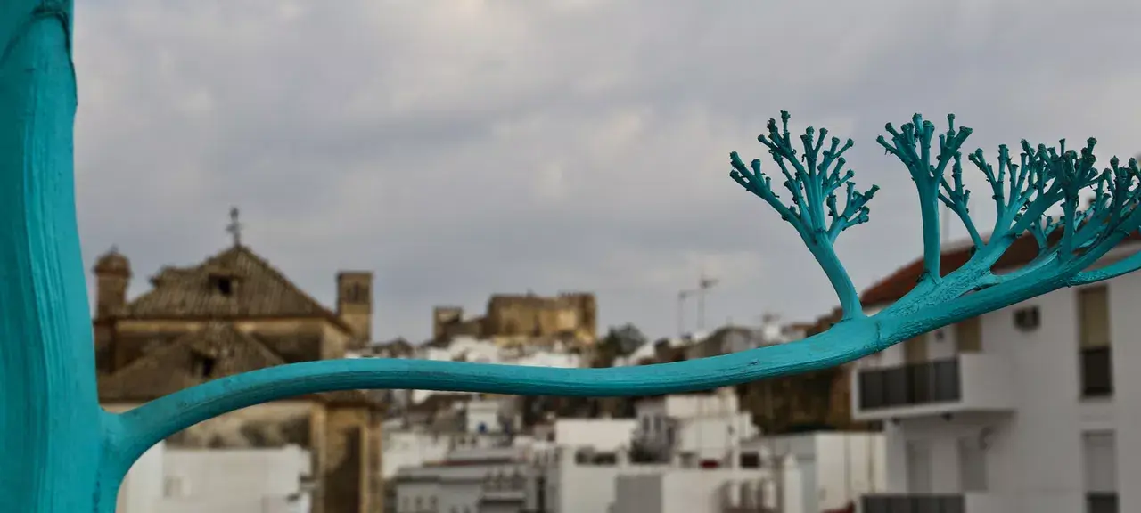 Escultura de árbol azul en primer plano con vista a un pueblo con edificios blancos y una iglesia al fondo bajo