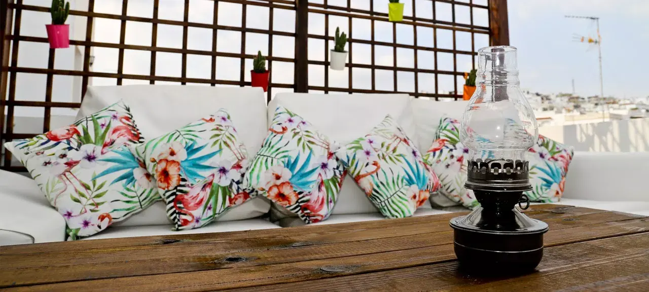 Sofá exterior con cojines de estampado floral y farol de aceite sobre mesa de madera, con celosía y macetas al fondo.