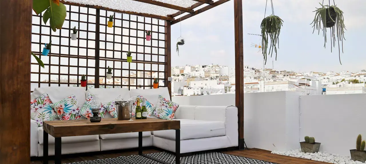 Terraza exterior con sofá blanco en L, mesa de centro de madera, cojines con estampado tropical, celosía de madera