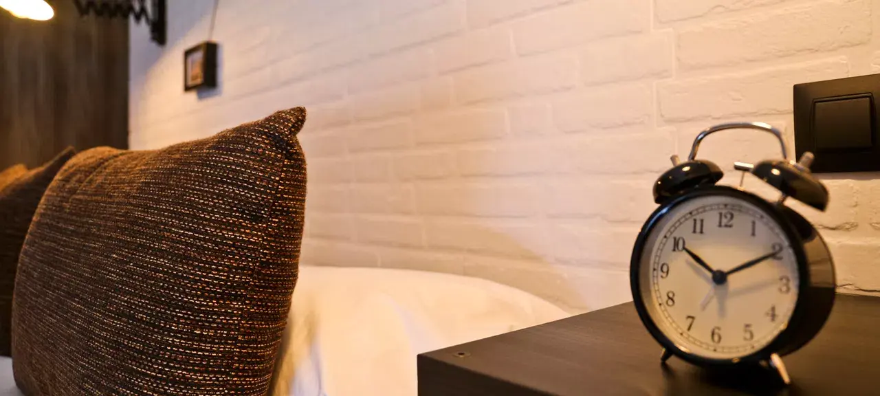 Detalle de una habitación con una almohada texturizada en tonos marrones sobre una cama, un reloj despertador negro