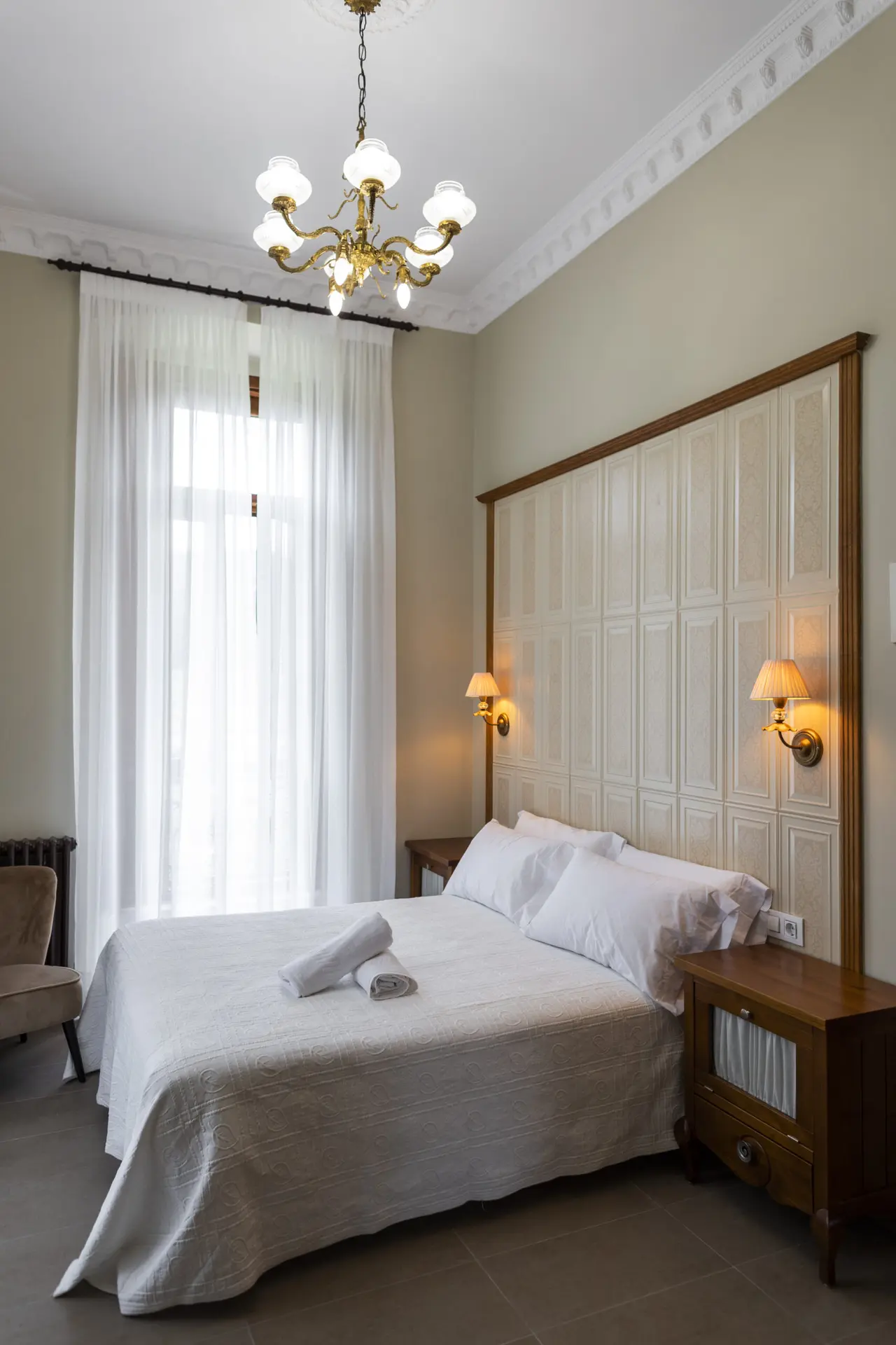 Habitación con cama doble vestida de blanco, cabecero alto de paneles, dos lámparas de pared, mesita de noche