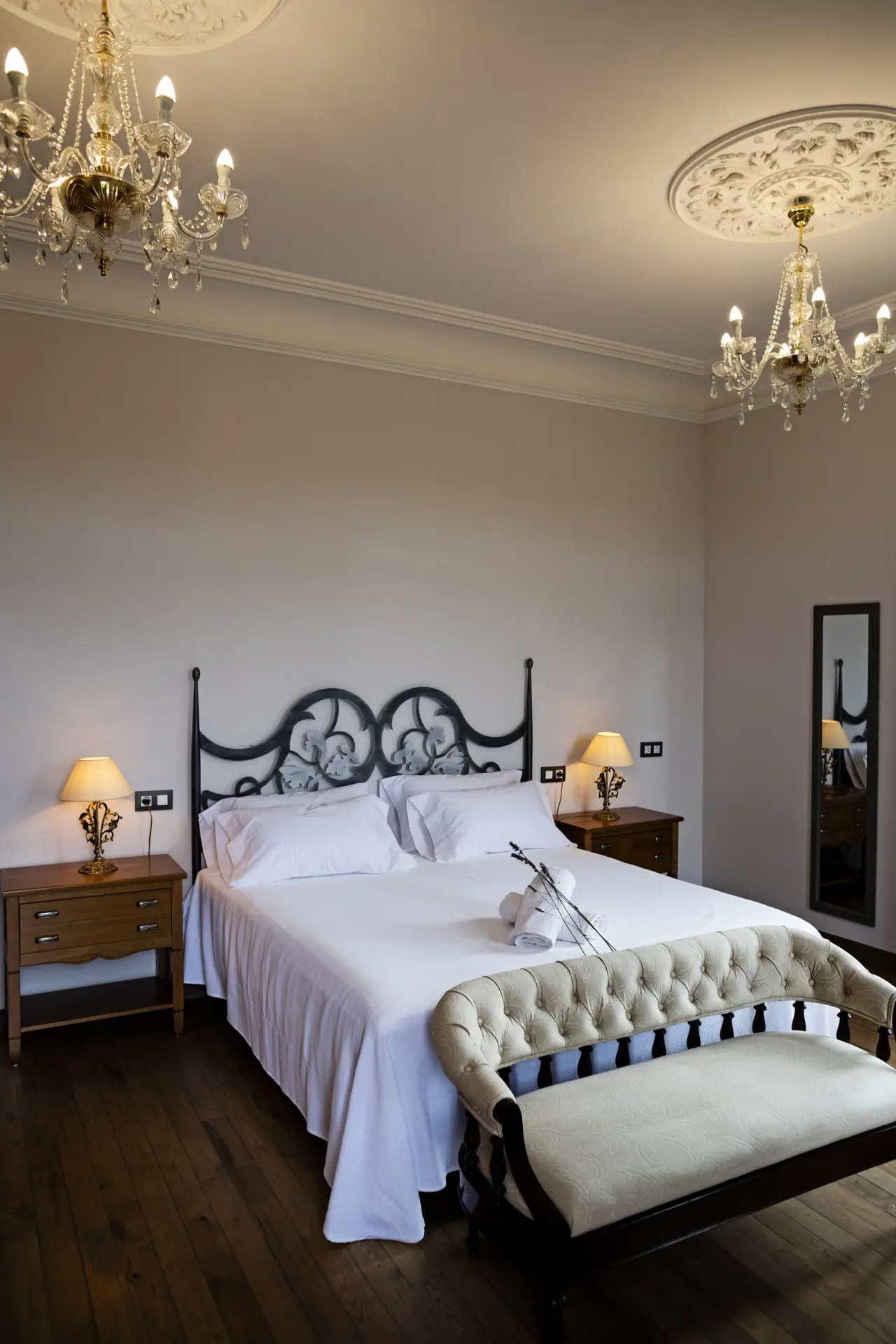 Habitación con cama doble de matrimonio con cabecero de forja negro, dos mesitas de noche con lámparas, banco tapizado