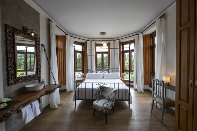 Vista de una suite con cama de matrimonio de hierro forjado en el centro, rodeada de ventanas con cortinas blancas