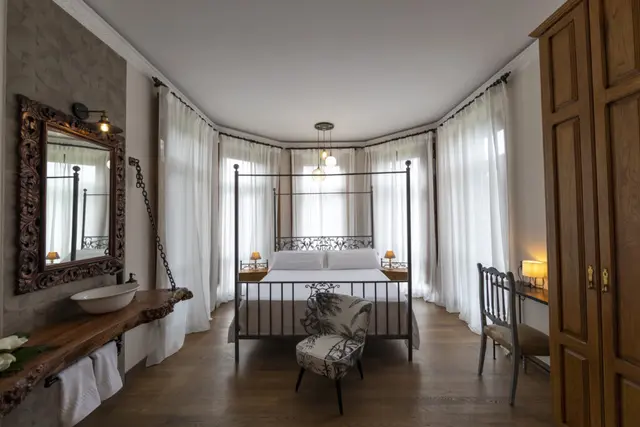 Vista de una suite con cama de matrimonio de hierro forjado en el centro, rodeada de cortinas blancas.