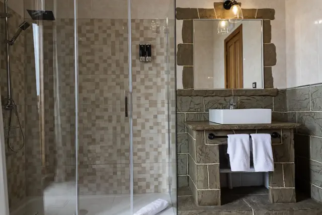 Baño con ducha de vidrio transparente a la izquierda y lavabo de piedra con espejo y toallas blancas a la derecha.